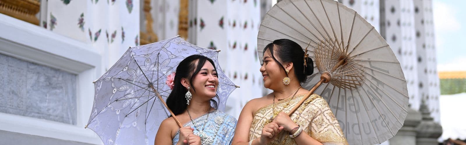 Two students wearing traditional Thai dresses and holding umbrellas walk next to a black and white temple